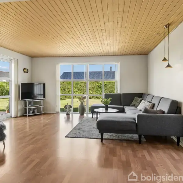 En moderne stue med en grå hjørnesofa og sofabord. En fladskærm står ved en stor glasdør, der leder ud til en have med græsplæne. Væggene er lyse, og loftet har træpaneler.