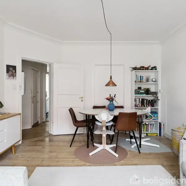 Et spisebord med stole står i et lyst rum med hvidmalede vægge og trægulv. En lampe hænger over bordet. Reol med bøger og legetøj i baggrunden. Ingen synlig tekst.