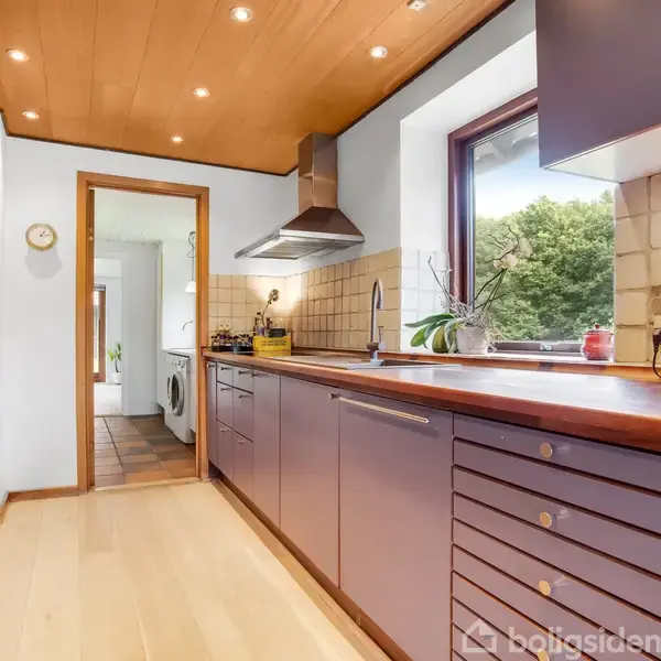 Køkken med træloft og lyse trægulve, står med udsigt til en terrasse gennem store glasdøre. Hylder fyldt med bøger ses til venstre.