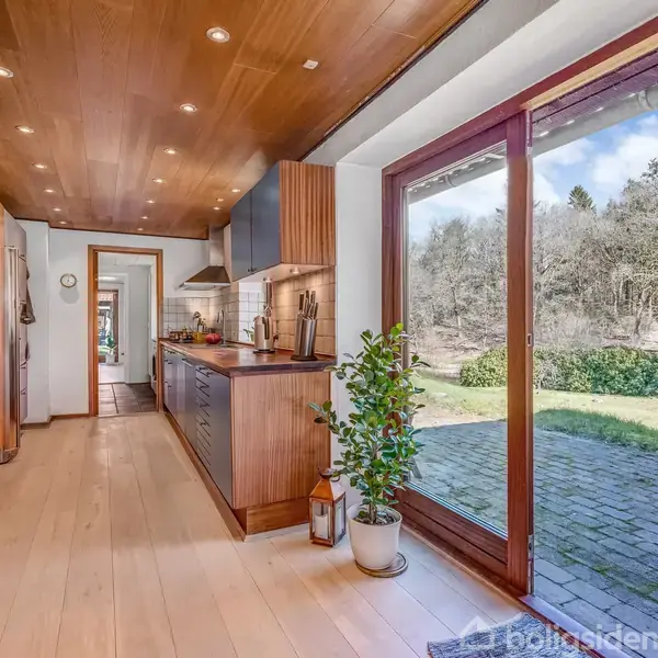 Spisestue med træbord og stole, lys rammer rummet gennem store vinduer; farverig lampe og maleri giver varme. Glasdør leder til tilstødende stue med flere træmøbler.