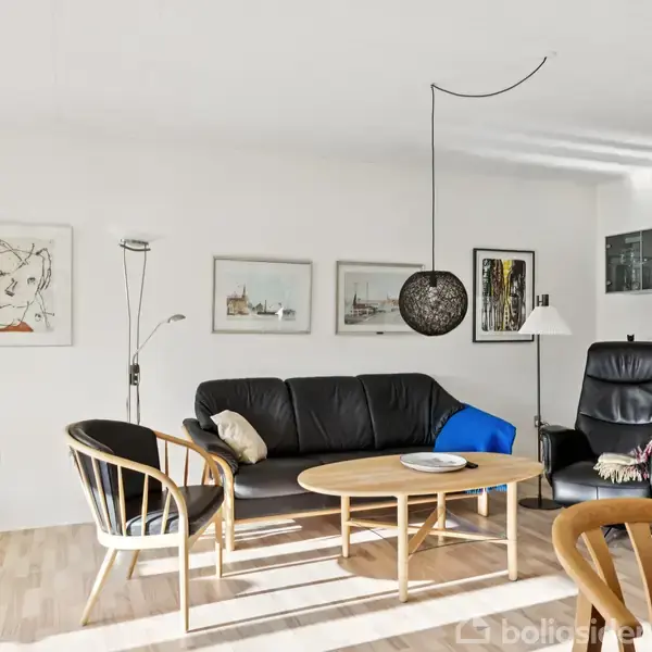 Sort sofa står i en stue med kunst på væggen, placeret mellem en gulvlampe og reol. Rundt sofabord og stole omringer sofaen. Grøn plante ved vinduet.