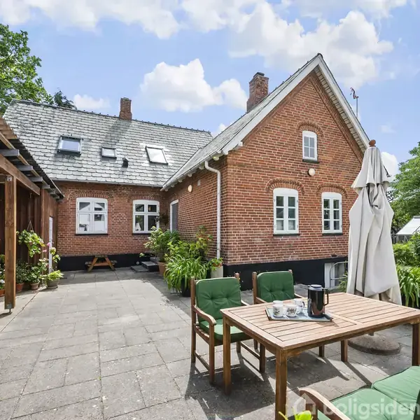 Rødt murstenshus med skrå tag, terrasse med bord og stole, parasol ved siden af. Haven omkranset af grønne buske og træer, drivhus i baggrunden. Klar, blå himmel over.