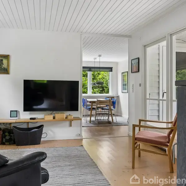 En stue med sort læderstol og fladskærms-TV på væggen. Et tilstødende spiseområde med spisebord og stole ses gennem en åben dør. Lyse vægge og trægulv.