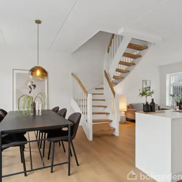 Et spisebord med seks stole står under en hængende lampe, mens en trappe fører opad i et moderne, lyst interiør med trægulve og minimalistisk indretning.