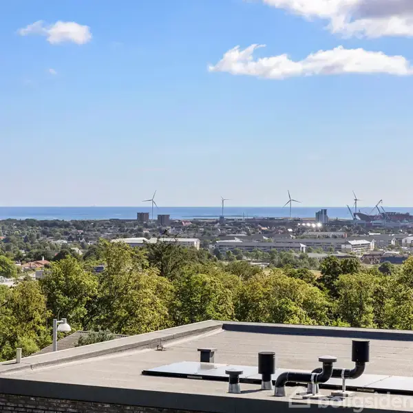 Fladt tag med ventilationsrør står stille under en klar blå himmel. I baggrunden ses et grønt skovområde, vindmøller og kystlinje ved en by.
