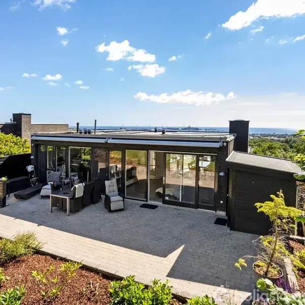 En moderne bungalow med store vinduer står på en terrasse, omgivet af en velplejet have med udsigt over en by i det fjerne under en klar blå himmel.