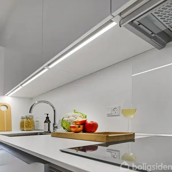 Køkkenbord med skærebræt, grøntsager og glas vin. Lys under overskabe oplyser området. Moderne indretning med indbygget kogeplade og vask. En krukke med pasta og en sæbedispenser står til venstre.