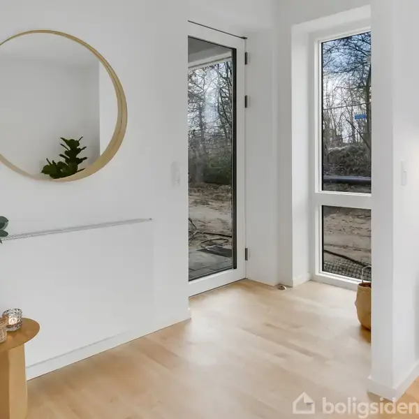 Et rundt spejl hænger på en hvid væg over et lille træbord med vase og lys. Døren ved siden af fører ud til en skovrig baggrund.