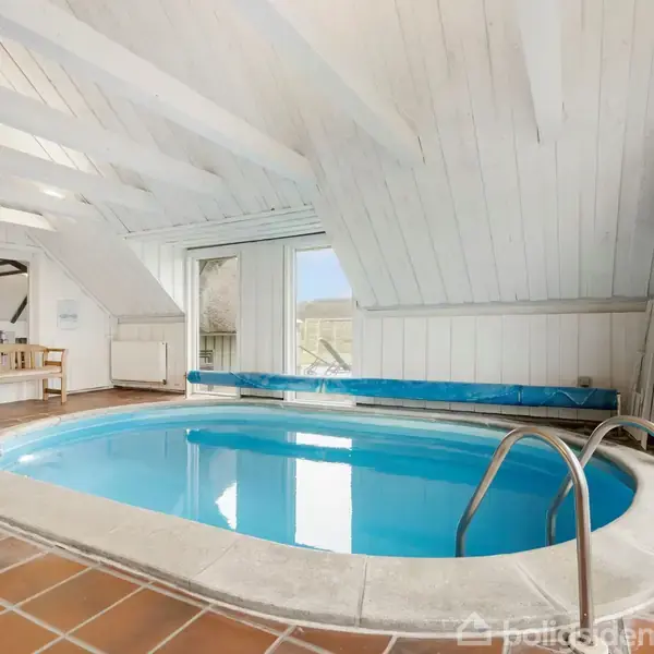 En indendørs swimmingpool med trapper fører ned i det blå vand. Poolen er omgivet af hvide trævægge og loft. Til venstre står et spisebord ved siden af vinduer med udsigt.