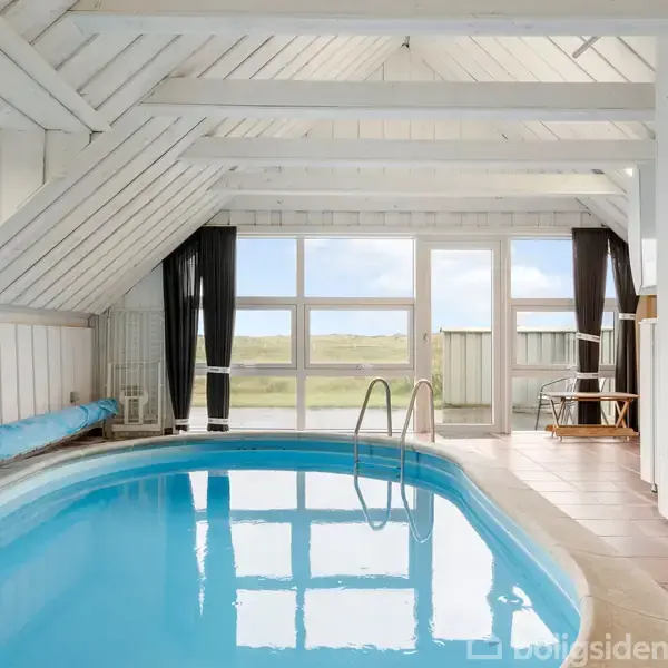 Indendørs swimmingpool med blå vand og stige, placeret i et lyst rum med hvide vægge og store vinduer, der giver udsigt til et grønt landskab. Møbler står langs siden.