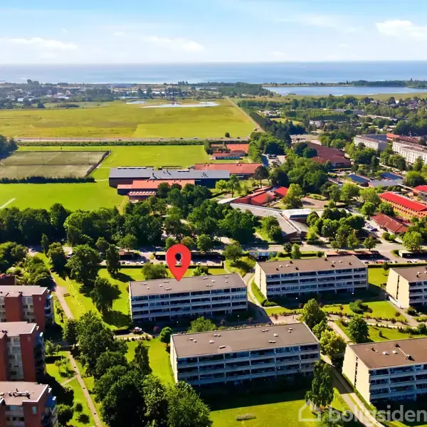 En række boligblokke strækker sig langs grønne områder med en rød markør over én bygning. Baggrund viser en kystlinje og skovklædte områder under en klar blå himmel.