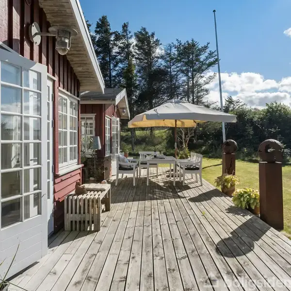 Rød træbygning med store vinduer har en træterrasse, hvor et bord- og stolearrangement står under en parasol. Terrassen er omringet af grønne buske og høje træer.