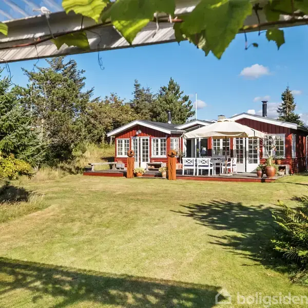 Rødt træhus med terrasse og havemøbler står i en frodig have omgivet af grønne buske og træer. Himmel med få skyer ses i baggrunden.