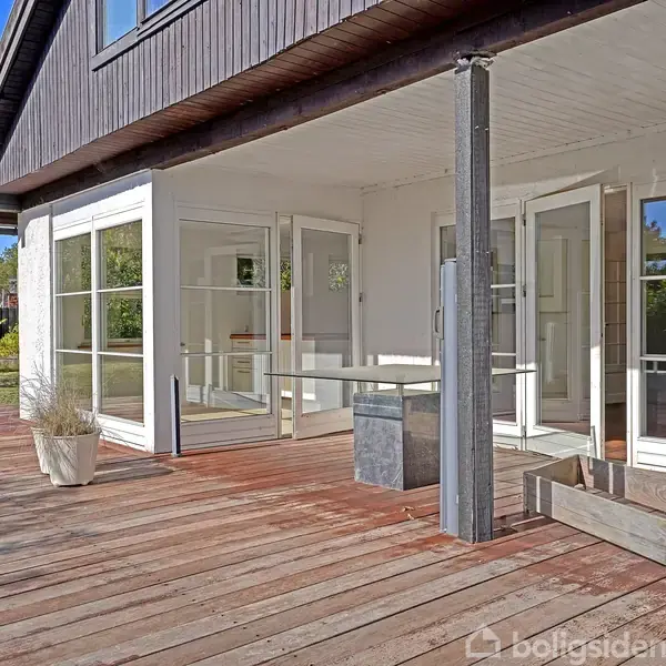 En træterrasse med krukker og plantekasser. Ved siden af er en moderne bygning med mange vinduer. Haven er delvist synlig med græsplæne og træer i baggrunden.