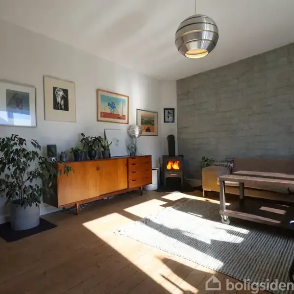 Stue med trækommode og billeder på væggen. Brændeovn i hjørnet afgiver varme. Lysegrå væg, gråbrun sofa, stort tæppe og grønne planter skaber hyggelig atmosfære.