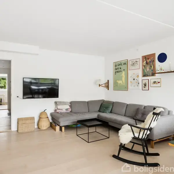 Grå sofa står i hjørnet af en stue, dekoreret med kunstværker og en hylde på væggen. Et fjernsyn hænger ved siden af en åben dør til en anden del af huset.
