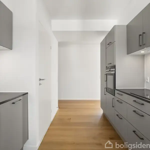 Moderne køkken med grå skabe og sorte bordplader; monteret med indbygget ovn og kogeplade. Trægulve strækker sig mod en lys døråbning i baggrunden.