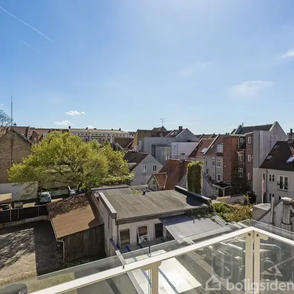 En byudsigt fra en altan viser tage af gamle murstenshuse under en klar blå himmel. En stor grøn trætop ses blandt bygningerne i baggrunden.