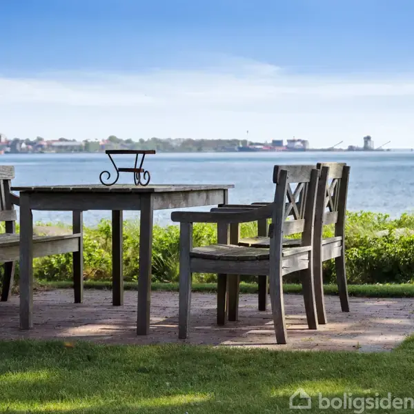 Træmøbler står på fliser nær en sø med udsigt over vandet og en by med kraner i baggrunden. Græs og buske omgiver området, og himlen er klar.