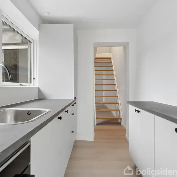 Et moderne køkken med hvide skabe og sort bordplade. En vask er monteret under et stort vindue. En åben døråbning viser en trappe i baggrunden.
