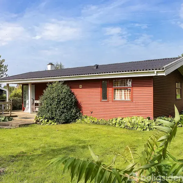 Et lille, rødt træhus står omgivet af en grøn have med buske og træer. Huset har hvide vinduer og en veranda med trækant. Himlen er blå og klar.