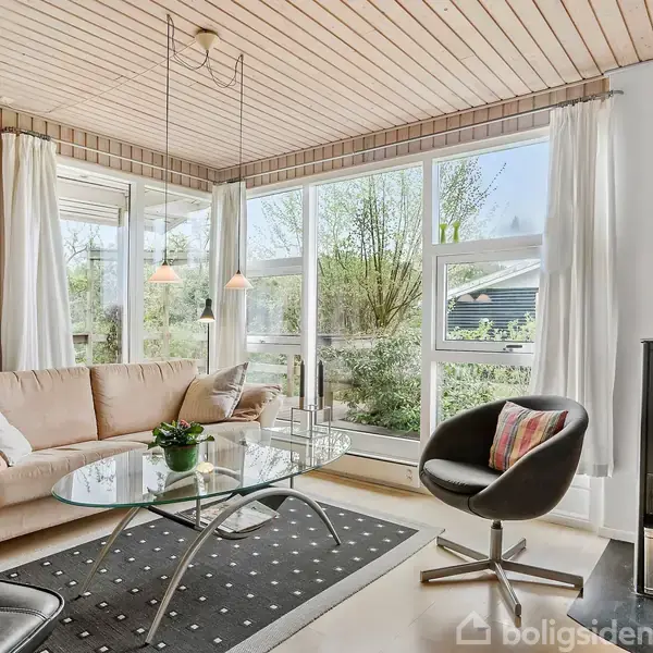 En stue med en beige sofa, pejs i hjørne, glasbord på tæppe og polstret stol. Store vinduer viser en have udenfor, og lys træbeklædning pryder loftet og vægge.