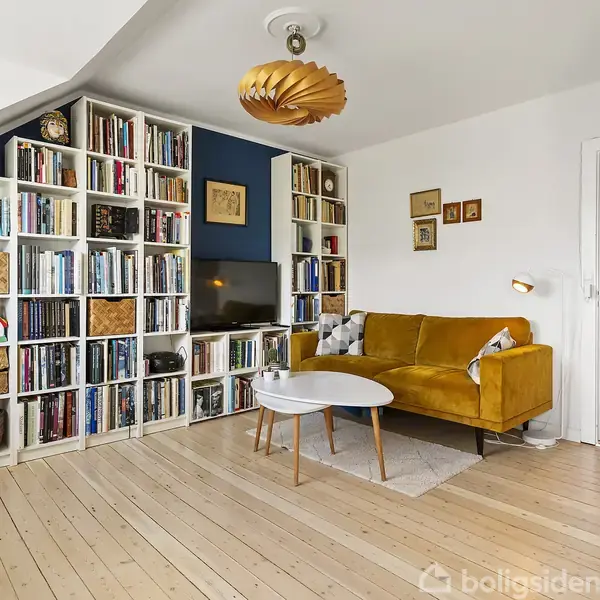 Stue med sofa i sennepsfarve, sofabord og en stor bogreol fyldt med bøger. Væggene er hvide, lyse trægulve, og en unik loftlampe hænger ned fra loftet.