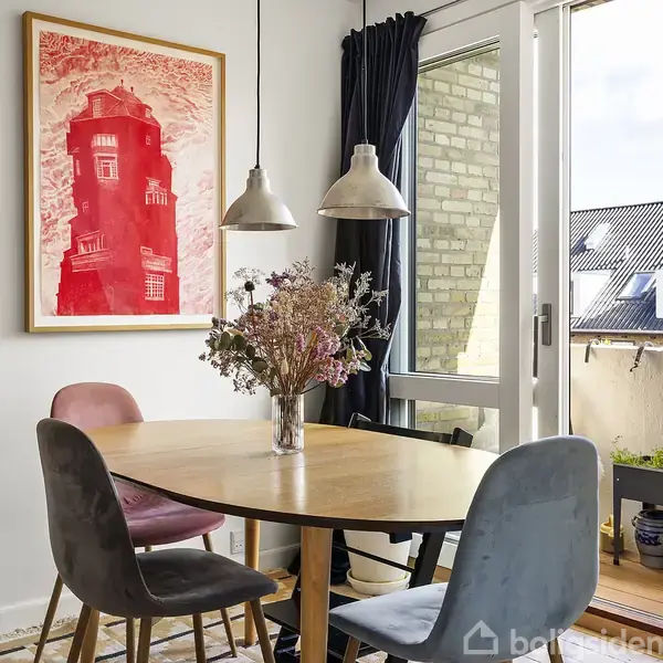 Et rundt træbord med fire forskellige stole står i et lyst rum. En vase med blomster er på bordet. På væggen hænger farverige plakater, og en glasdør fører til en altan.
