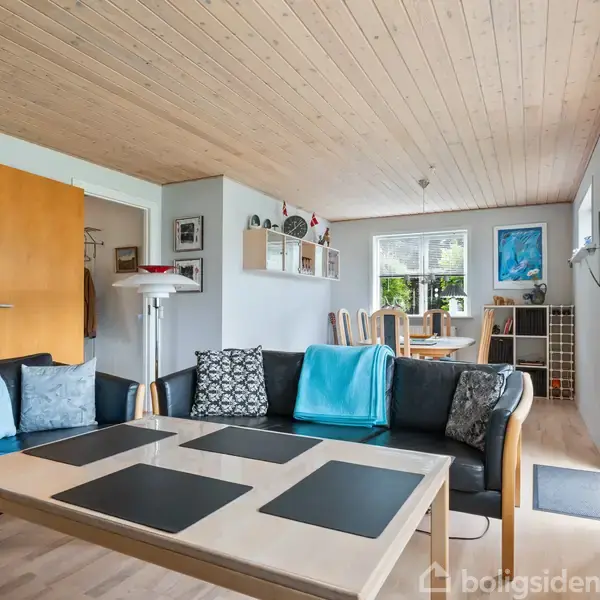 Stue med sort sofa og træspladebord, indrettet med puder og tæpper. I baggrunden står et spisebord i et lyst rum. Store vinduer til højre lader lys strømme ind.