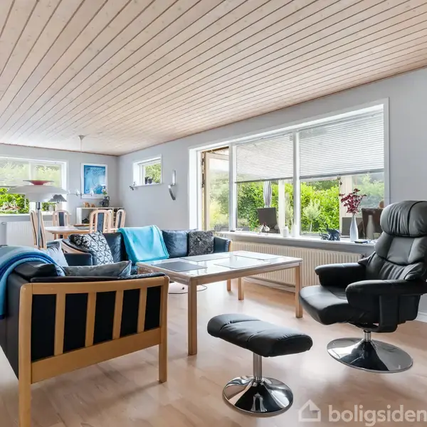 En stue med sort lænestol og fodskammel, sofa og træbord. Store vinduer viser grønne planter udenfor. Trægulv og loft, samt et spiseområde i baggrunden.