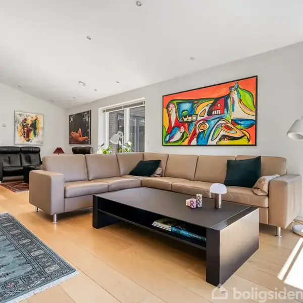En beige hjørnesofa er placeret i en moderne stue med lyse trægulve. Over sofaen hænger farverig kunst. Et sort sofabord står foran sofaen, omgivet af planter og naturligt lys.