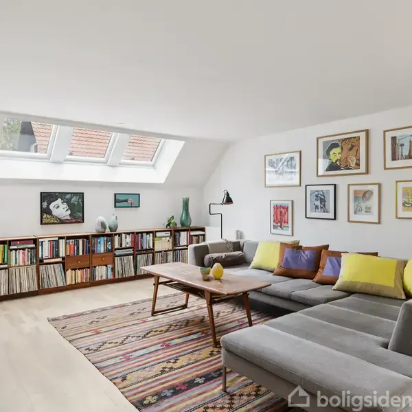 En lys stue med en grå, hjørnesofa fyldt med farverige puder. Sofaen står overfor et lavt træbord. Væggene har dekorative billeder, og der er en lang bogreol under skråvinduer.