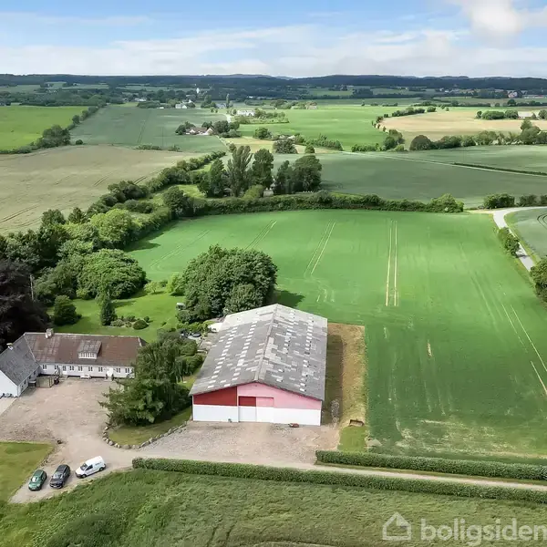 Bygning og lade står stille i et landligt område med omgivende marker og blå himmel. Mindre grusvej med biler foran bygningen, og grønne træer skaber en naturlig grænse.
