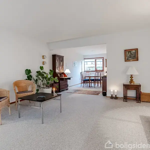 Stue med tæppebelagt gulv, kurvestole omkring et sofabord, planter ved siden af. Baggrund viser en spisestue gennem en bred åbning, med bord, stole og vindue.