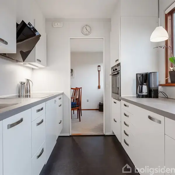 Et moderne køkken med hvide skabe og stålgreb strækker sig ind i en spisestue. En kaffemaskine står ved siden af planter på bordet under vinduet. Ingen tekst fundet.