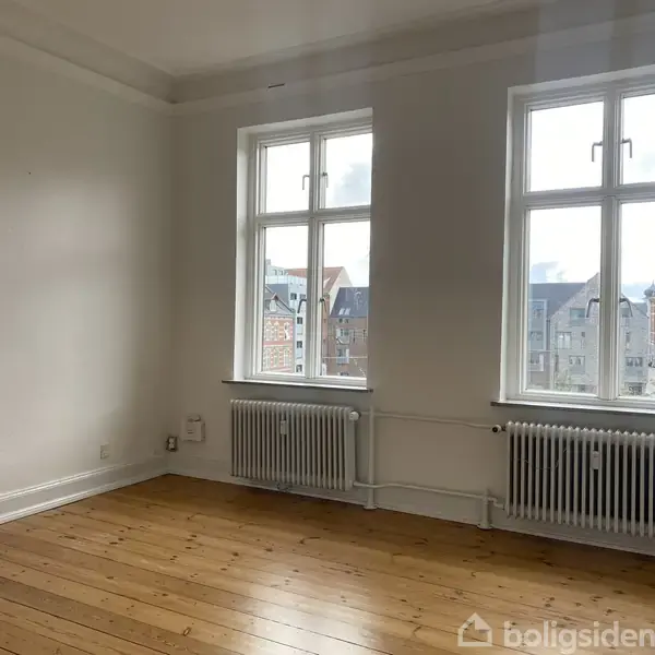 Et tomt værelse har trægulv og to store vinduer, som slipper lys ind. Radiatorer er placeret under vinduerne. Udenfor ses bygninger og himmel.