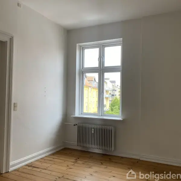 Et lyst, tomt rum med trægulv har et vindue åbent mod en bygning udenfor. Der står en radiator under vinduet, og en lukket dør ses til venstre.