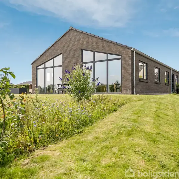 En moderne, en-etagers bygning med store vinduespartier står i en velplejet have. Græsset er nyslået, og forskellige planter vokser langs bygningens kant. Skyfri blå himmel i baggrunden.