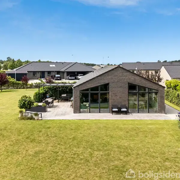 Moderne hus med store glaspartier står på en velplejet græsplæne. Terrassen er udstyret med udemøbler. Omkringliggende området har flere huse og grønne træer under en blå himmel.