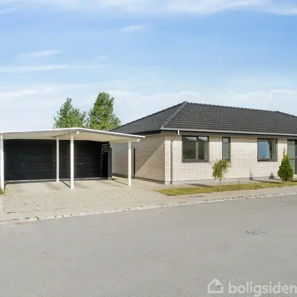 En enkelthus med moderne design og gråt tag står langs en stille villavej. Foran huset er der en garage med et fladt tag og en velplejet have med buske.