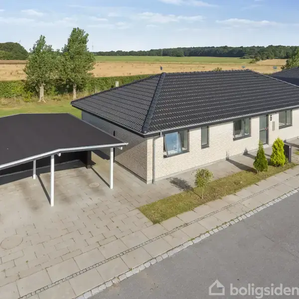 En enetages, moderne villa med sort tegltag står ved siden af en carport. Haven har græs og gyngestativ. Beliggende på en stille gade i landlige omgivelser med marker i baggrunden.