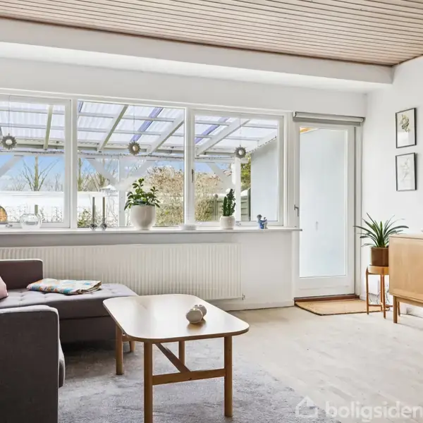 Gråt sofaarrangement med puder står i en lys stue med stort vindue og glasdør til en overdækket terrasse. En tv-skærm hænger på væggen over et træskab.