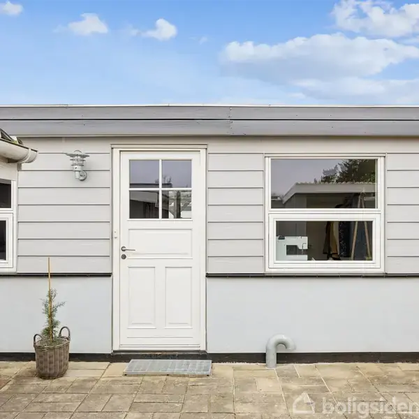 En hvid dør med vinduespanel er centreret i en gråmalet husfacade. Fliser ligger foran indgangen, og en lille plante i en kurv står ved døren.