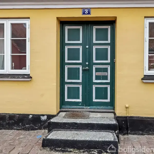 Grøn dobbeltdør står mellem to hvide vinduer i en gul murstensbygning. På dørens overligger er et blåt skilt med tallet 3. Trin fører op til døren.