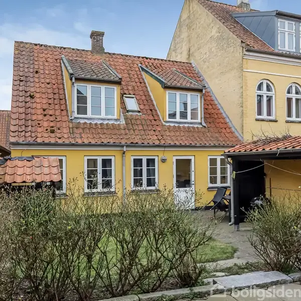 Gult murstenshus med rødt tegltag og små kviste; står stille i en gård omkranset af buske, med en sti foran og tilstødende bygning i baggrunden.