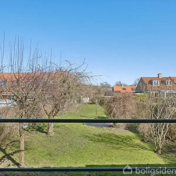 Balkonen har udsigt til en grøn have med træer og buske. Omkringliggende huse med røde tage kan ses i baggrunden under en klar blå himmel.
