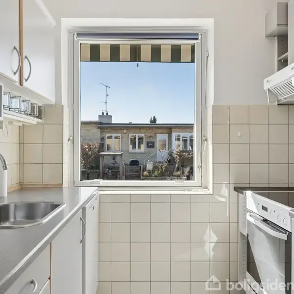 Et køkken med en moderne vask, komfur og opvaskemaskine, lyses op af et stort vindue. Udenfor ses et rækkehus og en klar himmel.
