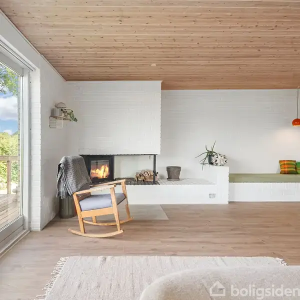 Et lyst, moderne stueinteriør med en gyngestol foran en tændt pejs. Stuen har store vinduer, en træloft og en grøn bænk med puder.