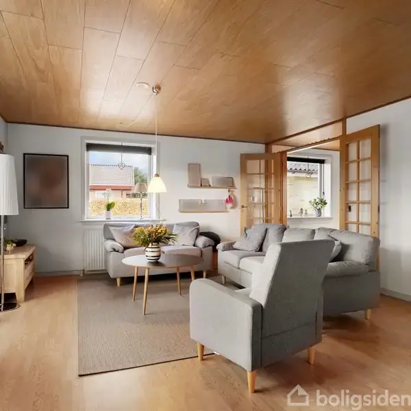 Stue med grå sofaer og et sofabord på et tæppe, omgivet af træhylder og glasdøre. Store vinduer slipper naturligt lys ind, og en træloft tilføjer varme.