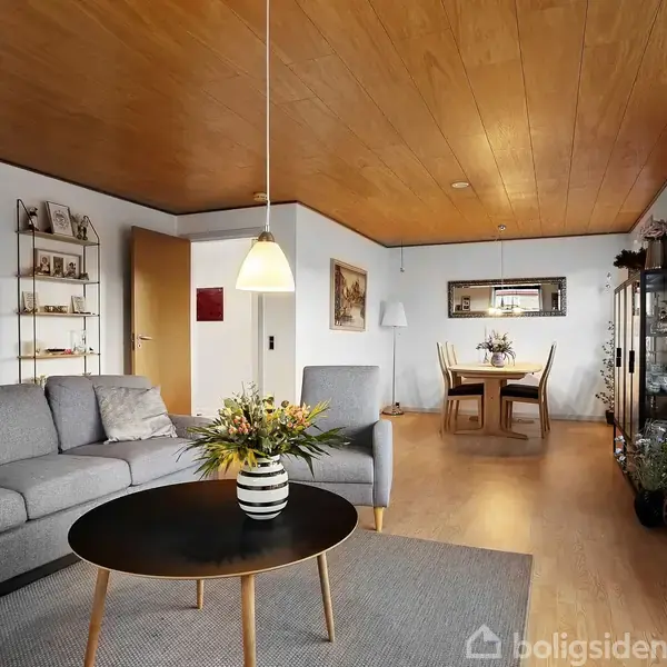 Grå sofa står i en stue med et rundt sofabord og en pendellampe ovenover. Stuen har trægulve, træloft og et spisebord i baggrunden ved et vindue.